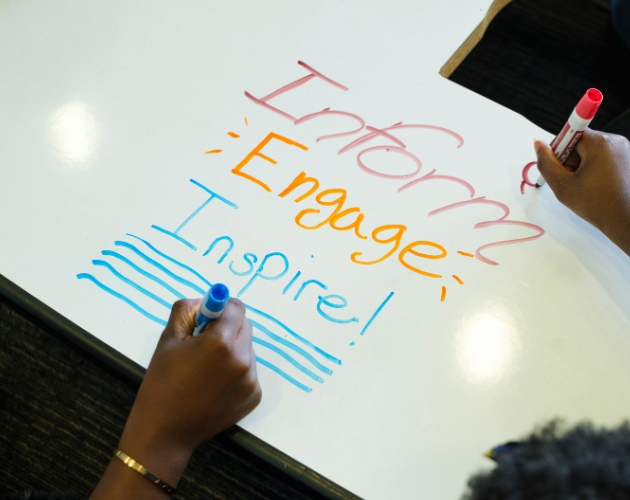Hands writing on a whiteboard with colorful marker strokes and lines, while another person holds a red marker near the top right.