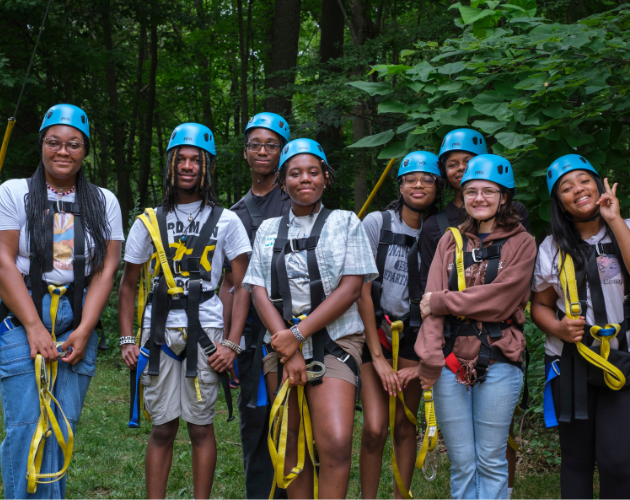 Group of people outdoors in a green forest wearing blue helmets and safety lanyards, posing together for a photo in casual clothes.