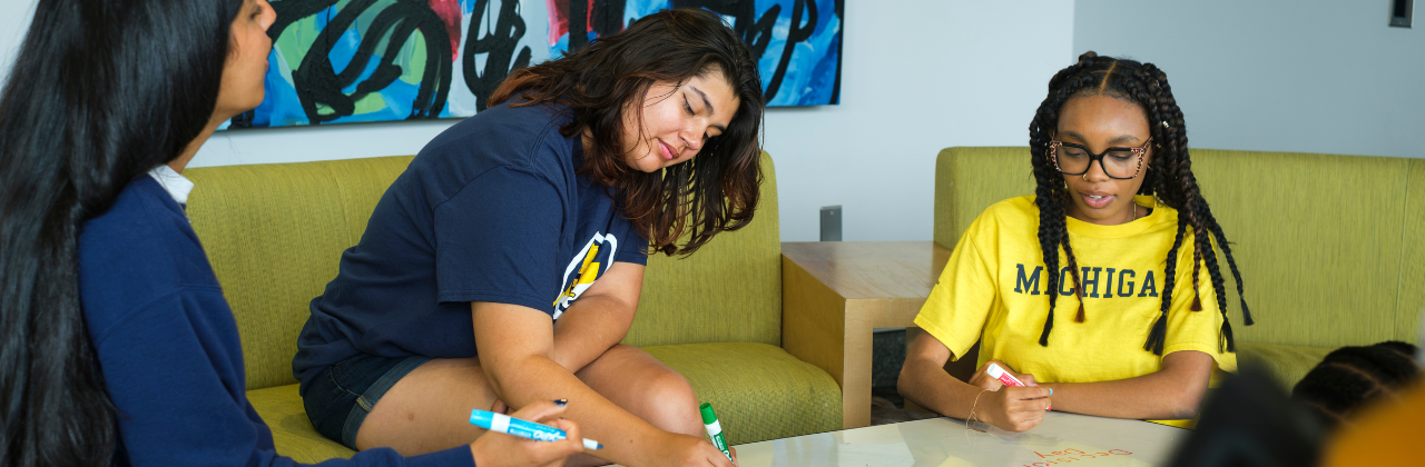 Three students sit around a table in a room with a colorful graffiti painting, each drawing with colored markers on paper.