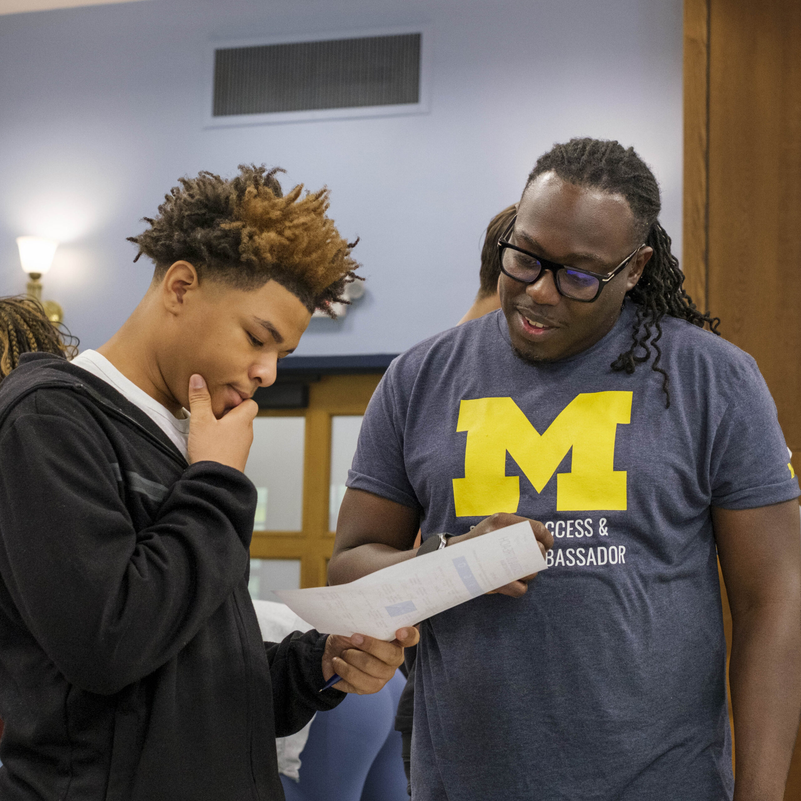 Two people in a classroom share a paper while talking; one wears a dark hoodie and the other a blue T-shirt with a large “M,” with other students standing and chatting in the background.